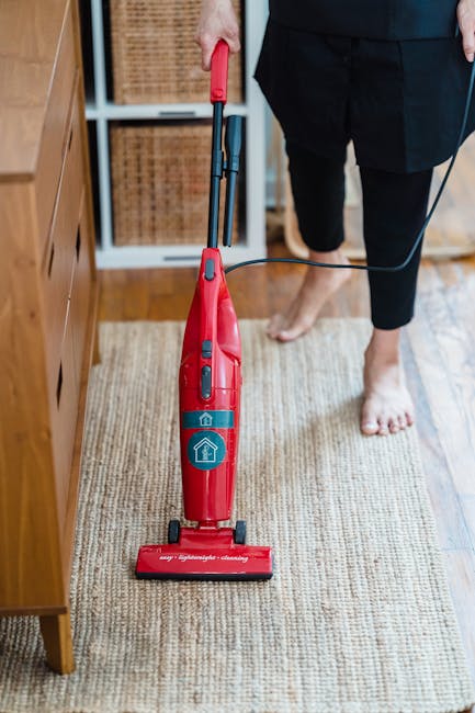 A person using a yellow vacuum cleaner with a black hose and a wide cleaning head to clean a patterned area rug in a room. The individual is wearing beige shoes, blue jeans, and a coat, and is standin